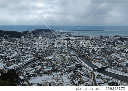 An aerial photograph showing a snowy landscape of Wajima City, Ishikawa Prefecture, with a view of the river mouth and the Sea of Japan. 139898175
