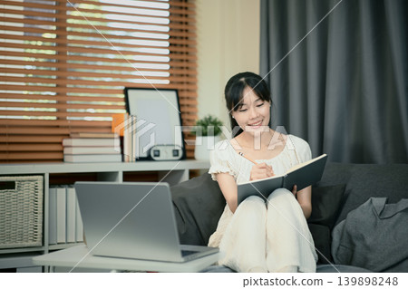 Smiling young woman remotely, sitting on sofa with notebooks and laptop, taking notes Smiling young woman remotely, sitting on sofa with notebooks and laptop, taking notes 139898248