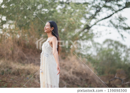 Woman in white lace dress standing in profile under green trees, breathing calmly in soft outdoor light, peaceful nature lifestyle, summer fashion, wellness, relaxation, freedom, and travel. Woman in white lace dress standing in profile under green trees, breathing calmly in soft outdoor light, peaceful nature lifestyle, summer fashion, wellness, relaxation, freedom, and travel. 139898300