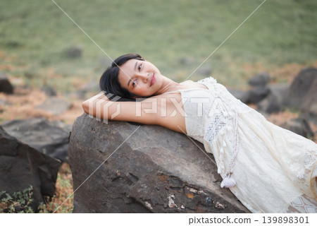 Woman in white lace dress resting on a rugged rock, gazing at camera in natural outdoor light, peaceful bohemian lifestyle, serene travel portrait, relaxation, wellness, summer fashion, and beauty. Woman in white lace dress resting on a rugged rock, gazing at camera in natural outdoor light, peaceful bohemian lifestyle, serene travel portrait, relaxation, wellness, summer fashion, and beauty. 139898301