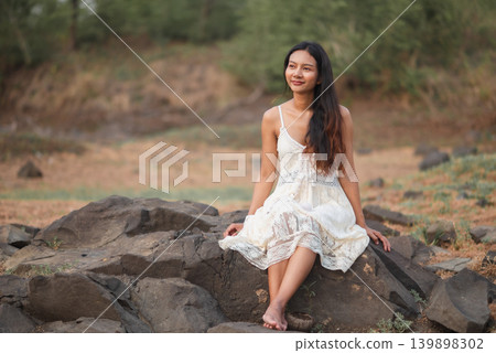 Woman in white lace dress sitting on rugged rocks in a quiet natural landscape, soft outdoor light, peaceful bohemian portrait, summer fashion, wellness lifestyle, travel, and serene relaxation. Woman in white lace dress sitting on rugged rocks in a quiet natural landscape, soft outdoor light, peaceful bohemian portrait, summer fashion, wellness lifestyle, travel, and serene relaxation. 139898302