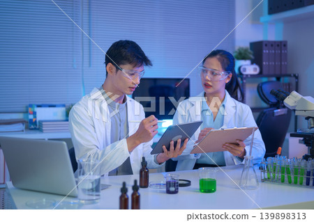 Two scientists discuss and record experimental data using tablet and clipboard in a modern laboratory with liquid samples and equipment Two scientists discuss and record experimental data using tablet and clipboard in a modern laboratory with liquid samples and equipment 139898313
