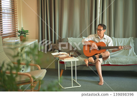 Man playing acoustic guitar on a sofa at home, wearing a prosthetic leg, relaxed leisure time and independent adaptive lifestyle. 139898396