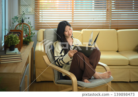 Happy relaxed young woman resting on armchair reading book and holding coffee cup Happy relaxed young woman resting on armchair reading book and holding coffee cup 139898473