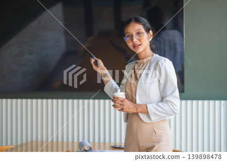Businesswoman in white blazer holding coffee and remote near office screen, presenting ideas in modern meeting room, showing confident speaker, corporate planning, and workplace communication. 139898478