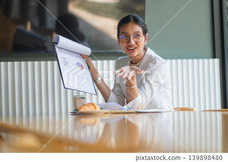 Businesswoman holding report charts and gesturing toward viewers in modern office, sharing financial insights, marketing results, strategy ideas, and confident teamwork presentation. 139898480