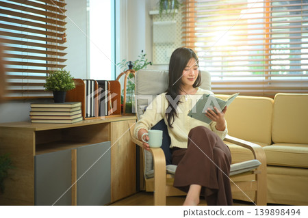 Happy relaxed young woman resting sitting on armchair reading daily and holding coffee cup Happy relaxed young woman resting sitting on armchair reading daily and holding coffee cup 139898494