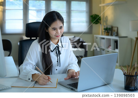 Smiling young Asian businesswoman or student working on a laptop and taking notes in a notebook, remote work or e-learning concept Smiling young Asian businesswoman or student working on a laptop and taking notes in a notebook, remote work or e-learning concept 139898503