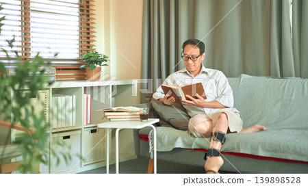 Middle-aged man reading a book on a sofa at home, wearing a prosthetic leg, calm leisure time and independent adaptive lifestyle. Middle-aged man reading a book on a sofa at home, wearing a prosthetic leg, calm leisure time and independent adaptive lifestyle. 139898528