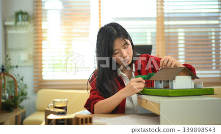Female architect examining house model at office desk, Professional architect reviewing architectural house model Female architect examining house model at office desk, Professional architect reviewing architectural house model 139898543