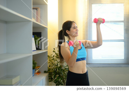Focused woman performing dumbbell shoulder exercise indoors in bright modern home environment Focused woman performing dumbbell shoulder exercise indoors in bright modern home environment 139898569