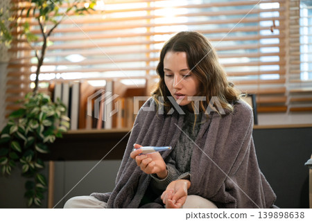 Woman wrapped in blanket checks temperature while resting indoors Woman wrapped in blanket checks temperature while resting indoors 139898638