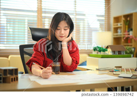 Female architect examining house model at office desk, Professional architect reviewing architectural house model Female architect examining house model at office desk, Professional architect reviewing architectural house model 139898742