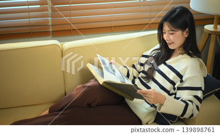 Happy relaxed young woman resting on sofa reading book and holding coffee cup Happy relaxed young woman resting on sofa reading book and holding coffee cup 139898761