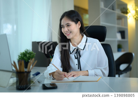 Smiling young Asian businesswoman or student working on a laptop and taking notes in a notebook, remote work or e-learning concept Smiling young Asian businesswoman or student working on a laptop and taking notes in a notebook, remote work or e-learning concept 139898805