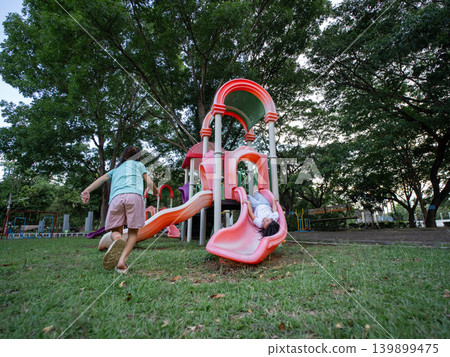 Two young Asian girls having fun playing on a colorful slide structure at the outdoor playground. 139899475