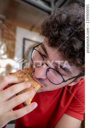New york, usa august 19,2024. Young man in glasses and red shirt bites into a delicious pizza slice in a manhattan pizzeria, savoring an urban meal moment 139900091