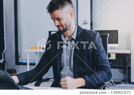 Smiling businessman in suit works on laptop in bright modern office Smiling businessman in suit works on laptop in bright modern office 139901075