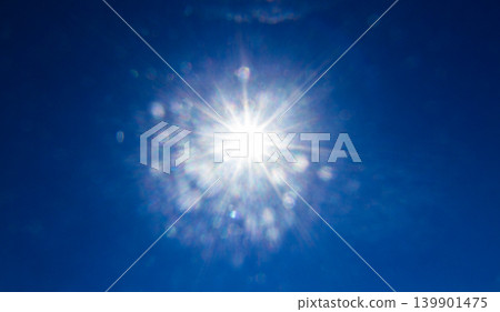 Sun shining brightly in a clear, deep blue sky, featuring a strong starburst effect with visible lens flare artifacts Sun shining brightly in a clear, deep blue sky, featuring a strong starburst effect with visible lens flare artifacts 139901475