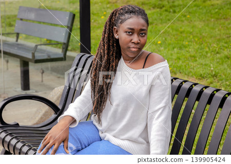 Young African woman sitting on bench at bus stop, urban park lifestyle 139905424