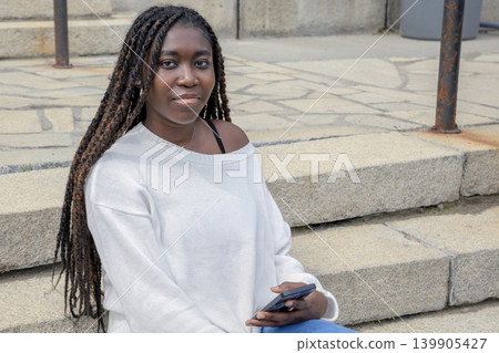 Young African student sitting on steps with smartphone, urban campus Young African student sitting on steps with smartphone, urban campus 139905427