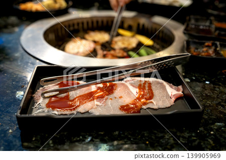 Fresh raw sliced pork with red marinade on a tray with metal tongs. Background shows a traditional yakiniku barbecue grill with meat cooking in a restaurant. Fresh raw sliced pork with red marinade on a tray with metal tongs. Background shows a traditional yakiniku barbecue grill with meat cooking in a restaurant. 139905969