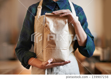 Barista woman, hands and bag while in an apron in background at home. Close up of female worker wearing pinafore and holding craft paper order while packaging coffee beans as small business owner Barista woman, hands and bag while in an apron in background at home. Close up of female worker wearing pinafore and holding craft paper order while packaging coffee beans as small business owner 139907238