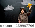 Child, boy and face by blackboard with plane for creative fun, playing or drawing of clouds for imagination. Student, kid and confidence with airplane, chalkboard or illustration for knowledge or sky 139907706
