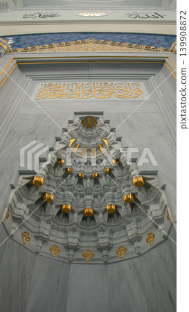 A close up of a marble mihrab shows detailed muqarnas designs and gold leaf accents. Soft natural light highlights the features in a mosque setting. A close up of a marble mihrab shows detailed muqarnas designs and gold leaf accents. Soft natural light highlights the features in a mosque setting. 139908872