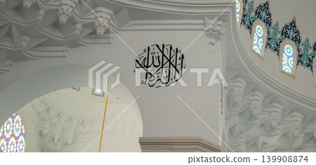A circular Arabic calligraphy mural is displayed on a clean white wall. The design is framed by traditional muqarnas architectural elements, adding historical context to the scene. A circular Arabic calligraphy mural is displayed on a clean white wall. The design is framed by traditional muqarnas architectural elements, adding historical context to the scene. 139908874