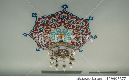 A detailed view shows a ceiling in a mosque, featuring a geometric star pattern in red and blue. A brass chandelier hangs below the ornate design. A detailed view shows a ceiling in a mosque, featuring a geometric star pattern in red and blue. A brass chandelier hangs below the ornate design. 139908877