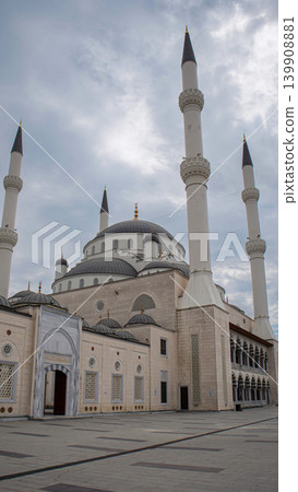 The scene shows a large mosque complex with tall minarets and a wide courtyard. People walk around the area while clouds cover the sky. The architecture reflects historical design. The scene shows a large mosque complex with tall minarets and a wide courtyard. People walk around the area while clouds cover the sky. The architecture reflects historical design. 139908881
