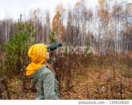 Woman birdwatcher looking through binoculars in autumn forest. Ecologist exploring nature and wildlife during fall season hike. Outdoor activity, adventure, travel, discovery and research concept. Woman birdwatcher looking through binoculars in autumn forest. Ecologist exploring nature and wildlife during fall season hike. Outdoor activity, adventure, travel, discovery and research concept. 139910689