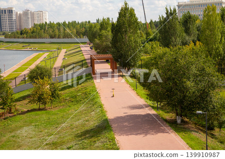 Yellow electric scooter parked on a paved path in a city park. Scenic summer landscape with green trees, lake embankment and modern architecture in background. Urban leisure concept. Yellow electric scooter parked on a paved path in a city park. Scenic summer landscape with green trees, lake embankment and modern architecture in background. Urban leisure concept. 139910987