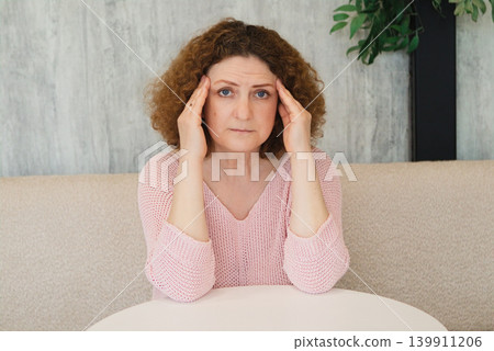 Woman holding her temples with headache and stress while sitting at a table in a living room Woman holding her temples with headache and stress while sitting at a table in a living room 139911206