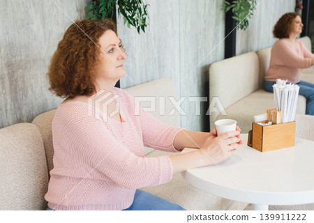 Middle-aged woman sitting in a cafe holding a coffee cup and looking thoughtful and relaxed Middle-aged woman sitting in a cafe holding a coffee cup and looking thoughtful and relaxed 139911222