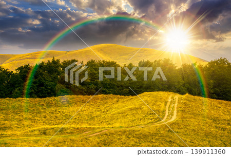 sunset over dirt road through grassy hills. mountain landscape with beech forest along the path. remote countryside. late summer scenery with primeval woods on hillside. weather with rainbow 139911360