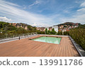 A modern rooftop terrace with a swimming pool. The area features wooden decking and overlooks a scenic view of buildings and hills under a clear blue sky. 139911786