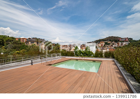 A modern rooftop terrace with a swimming pool. The area features wooden decking and overlooks a scenic view of buildings and hills under a clear blue sky. A modern rooftop terrace with a swimming pool. The area features wooden decking and overlooks a scenic view of buildings and hills under a clear blue sky. 139911786