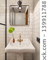 A modern bathroom with a white sink, gold faucet, and a green plant in a brown vase. The walls are tiled with white subway tiles and a mirror is mounted above the sink. 139911788