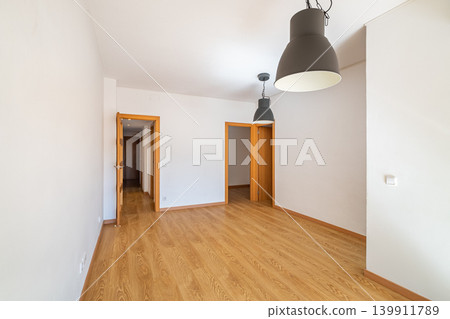 Empty room with wooden floor, white walls, and two pendant lights. Doors lead to other spaces. Bright and spacious atmosphere. Empty room with wooden floor, white walls, and two pendant lights. Doors lead to other spaces. Bright and spacious atmosphere. 139911789