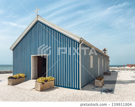 Perspective view of the iconic blue and white striped chapel of Praia de Mira nestled against the Atlantic ocean under a beautiful clear blue sky 139911804