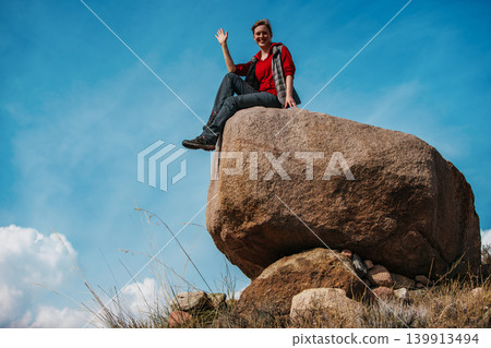 Young woman tourist sits on a big boulder and waves her hand Young woman tourist sits on a big boulder and waves her hand 139913494
