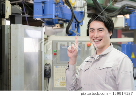 A young engineer smiles and points, making a pointing gesture, in a factory manufacturing area, performing quality control and equipment inspections. He is looking directly at the camera. A young engineer smiles and points, making a pointing gesture, in a factory manufacturing area, performing quality control and equipment inspections. He is looking directly at the camera. 139914187