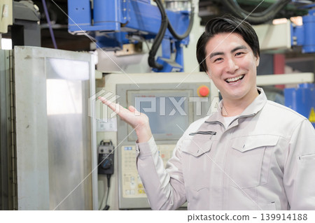 A young engineer in a factory production area smiles and points, showing his recommended pose for quality control and equipment inspection, looking directly at the camera. A young engineer in a factory production area smiles and points, showing his recommended pose for quality control and equipment inspection, looking directly at the camera. 139914188