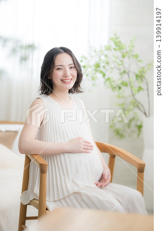 A Japanese pregnant woman smiles and places her hand on her stomach, sitting with a smile and looking directly at the camera in a maternity lifestyle image. A Japanese pregnant woman smiles and places her hand on her stomach, sitting with a smile and looking directly at the camera in a maternity lifestyle image. 139914197