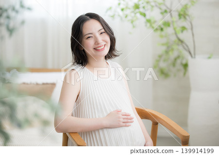 A Japanese pregnant woman smiles and places her hand on her stomach, sitting with a smile and looking directly at the camera in a maternity lifestyle image. A Japanese pregnant woman smiles and places her hand on her stomach, sitting with a smile and looking directly at the camera in a maternity lifestyle image. 139914199