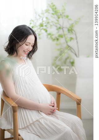 A Japanese pregnant woman with a smile and her hand on her stomach, a maternity lifestyle image with a downcast smile. A Japanese pregnant woman with a smile and her hand on her stomach, a maternity lifestyle image with a downcast smile. 139914206