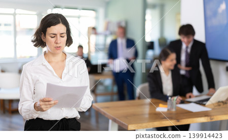 Positive young business woman with documents and pen in hand standing against background of colleague working in corporate office 139914423