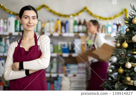 Young woman in apron stands among shelves and storefronts in household store, colleague in background Young woman in apron stands among shelves and storefronts in household store, colleague in background 139914827